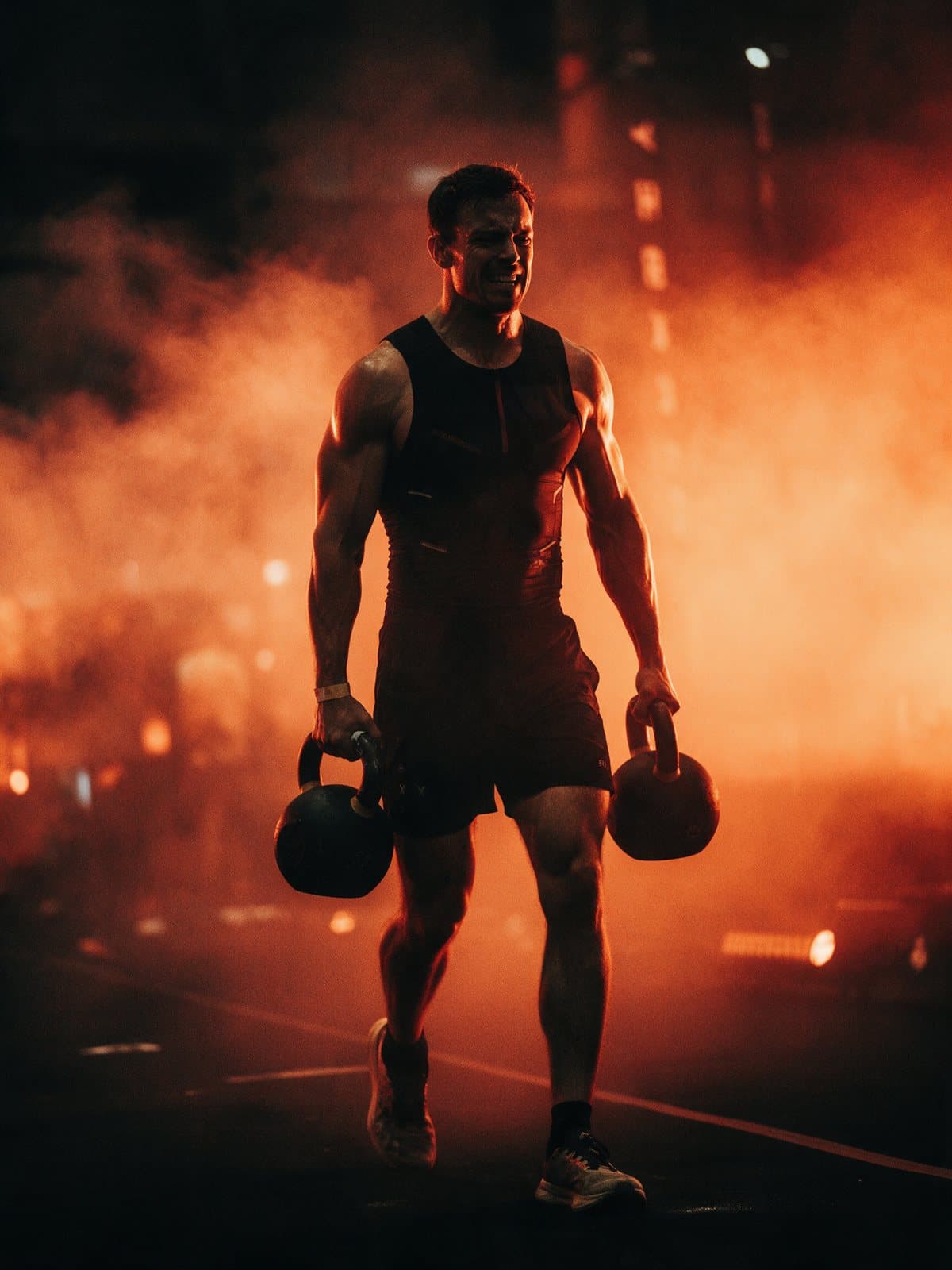 Hybrid athlete in race kit doing a kettlebell farmers carry through orange haze